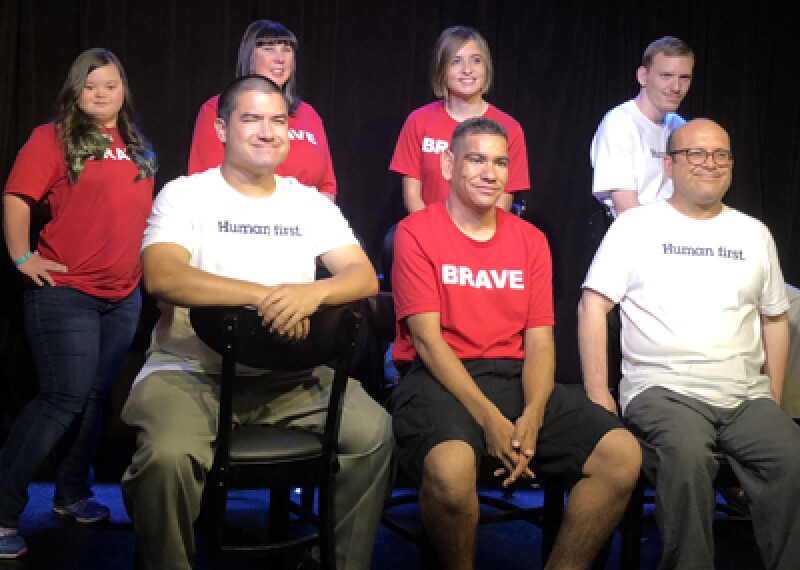 Special Olympics athletes in a group in front of a black backdrop. Athletes have on prinkshop t-shirts. Red shirts read, Brave; white shirts read, Human First. 