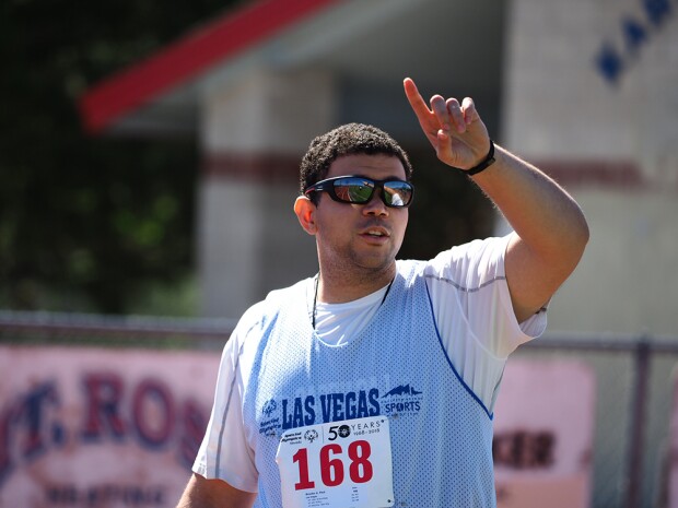 Young adult male on the field with one hand up and pointing his finger up to the sky. He has on sunglasses and a jersey that reads Special Olympics Las Vegas. 