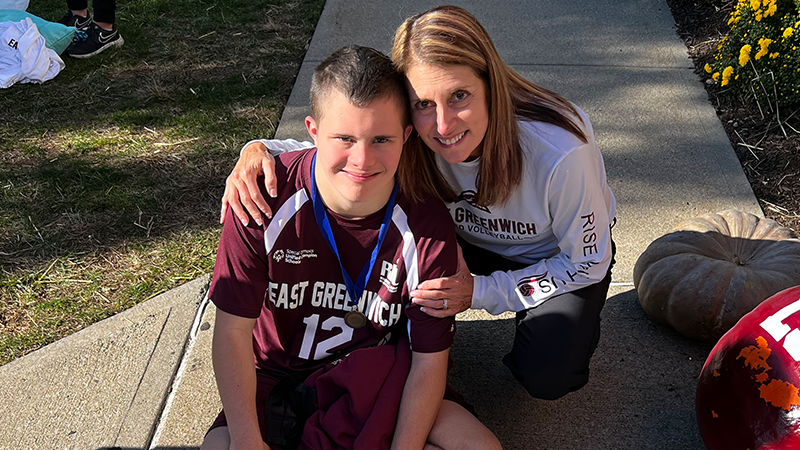 A Special Olympics athlete and his coach pose for a photo. They are both sitting on the ground and the coach has her arms around the athlete. The athlete is wearing a medal around his neck. 