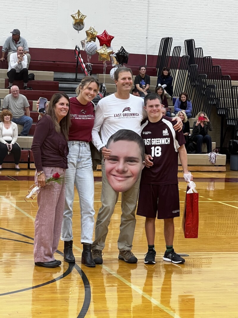 Four people (mom, sister, dad and brother) are standing on a basketball court posing for a family photo. The brother on the far right side has Down syndrome and is wearing a volleyball uniform. 