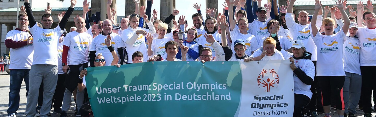 a large group of athletes and representatives cheering with their hands up and some are holding a banner in front of them. 